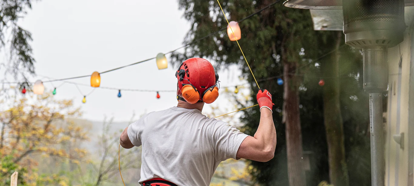 Arbeiter mit Schutzhelm und Ohrenschützern, der Lichterketten im Freien anbringt.