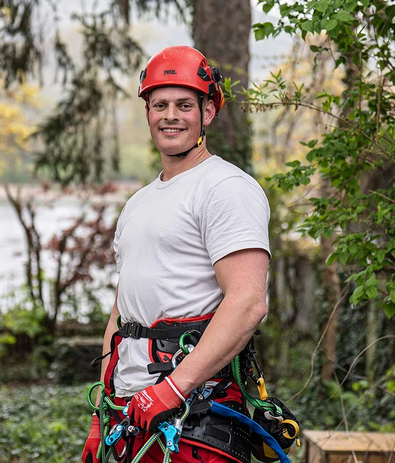 Ein Mann in Kletterausrüstung und Helm lächelt in der Natur, bereit für eine Baumkletteraktion.